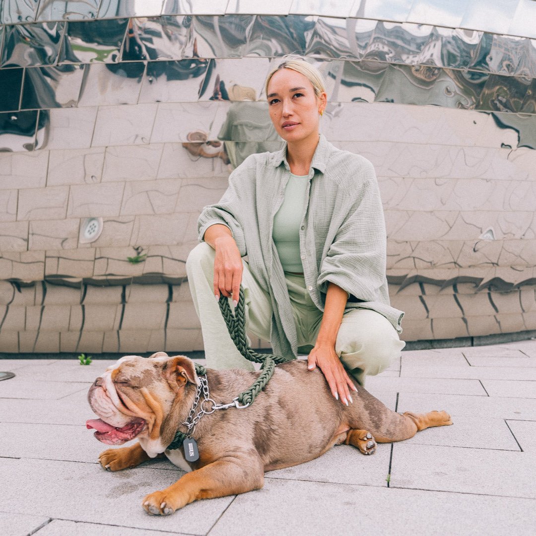 Frau mit olivgrüner Kleidung hält einen kräftigen Bulldoggen an der olivgrünen Power-Leash von Strong Dogs U.S., sitzend auf modernem Stadtpflaster vor spiegelnder Architektur.