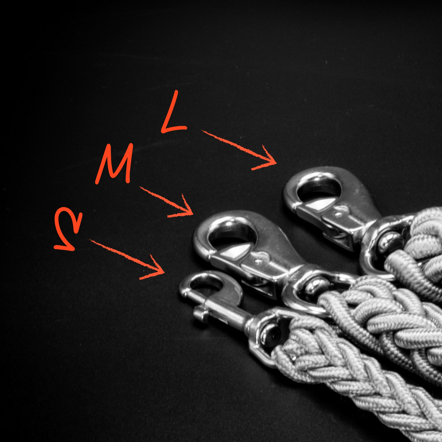 Größenvergleich der Power-Leash Karabiner von Strong Dogs U.S. in den Varianten S, M und L – drei geflochtene Leinen mit unterschiedlich großen Verschlüssen auf schwarzem Hintergrund, markiert mit roten Pfeilen und Beschriftung.